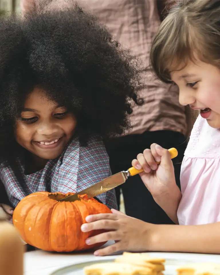 Two children carving a small orange pumpkin at a table—one smiling and holding the pumpkin steady while the other carefully cuts into the top with a kitchen knife.
